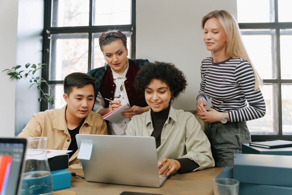 Coworkers discussing notes around a laptop in an office, illustrative stock photo